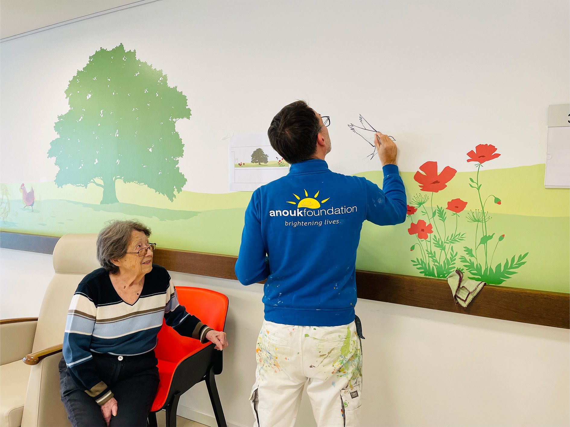 Two people painting a colorful mural on a hallway wall, with a seated person watching nearby.