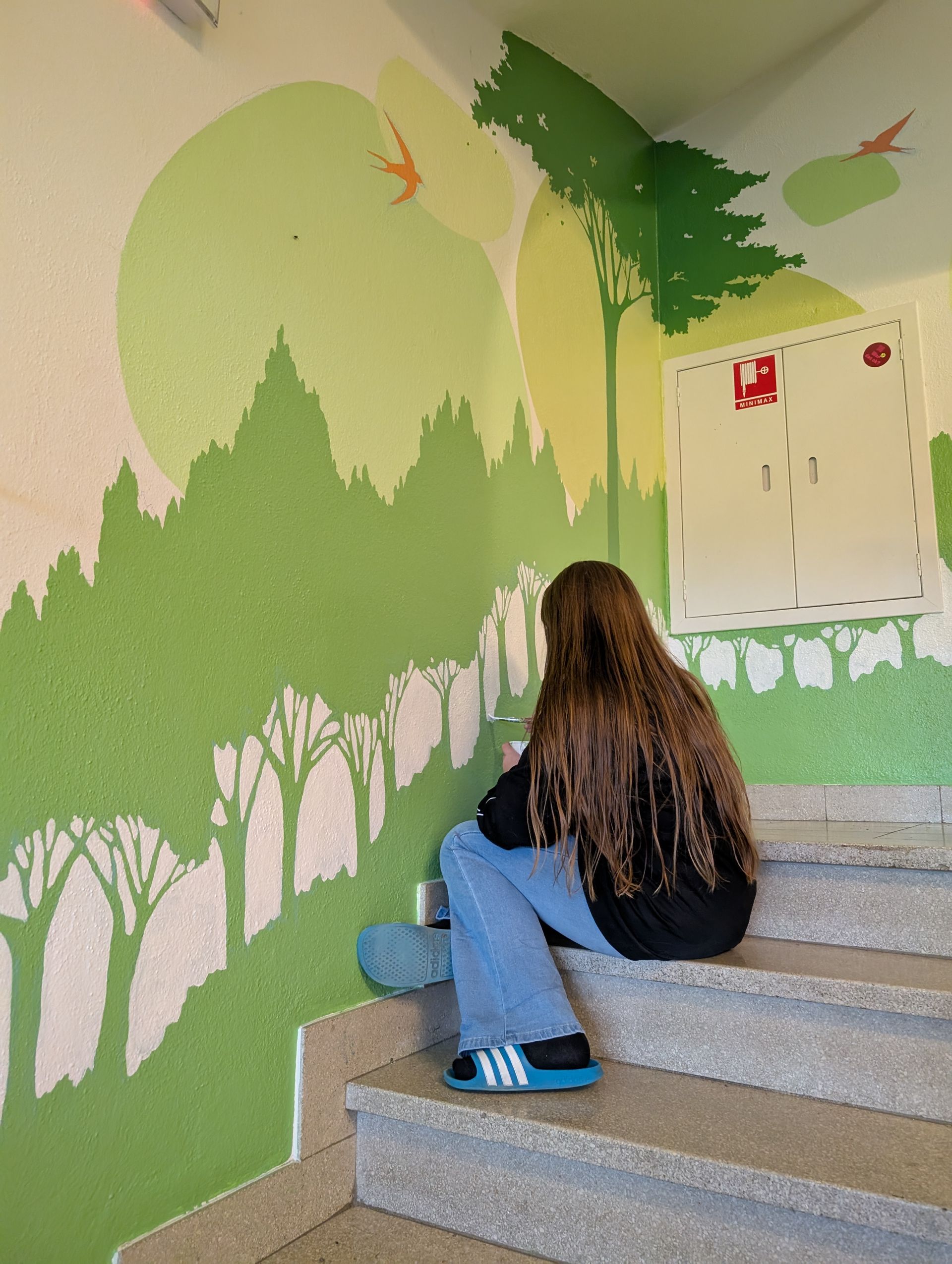 Person sitting on stairs beside a green tree mural in a stairwell