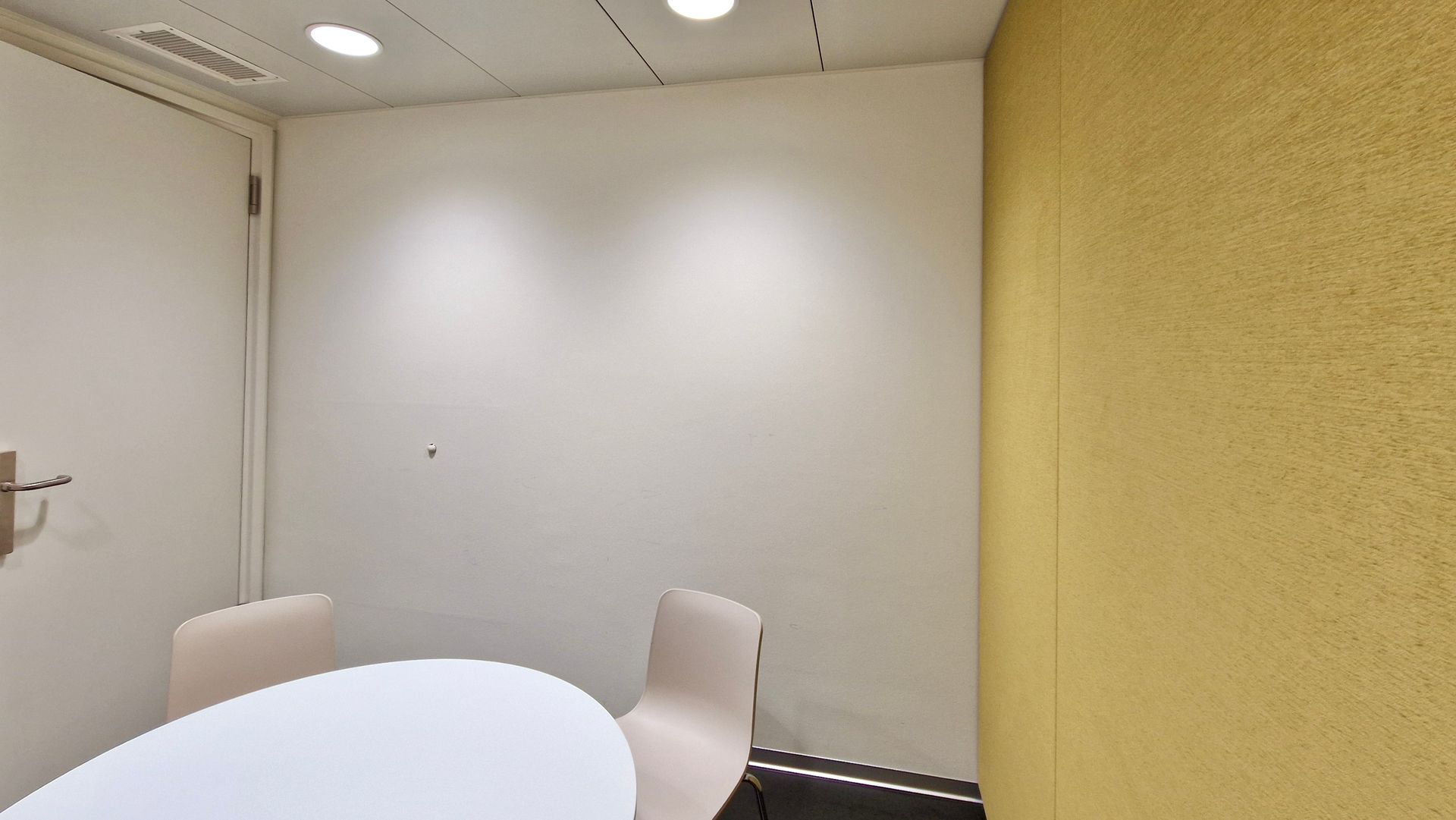 Small meeting room with a round white table, two beige chairs, and white walls beside a yellow partition.