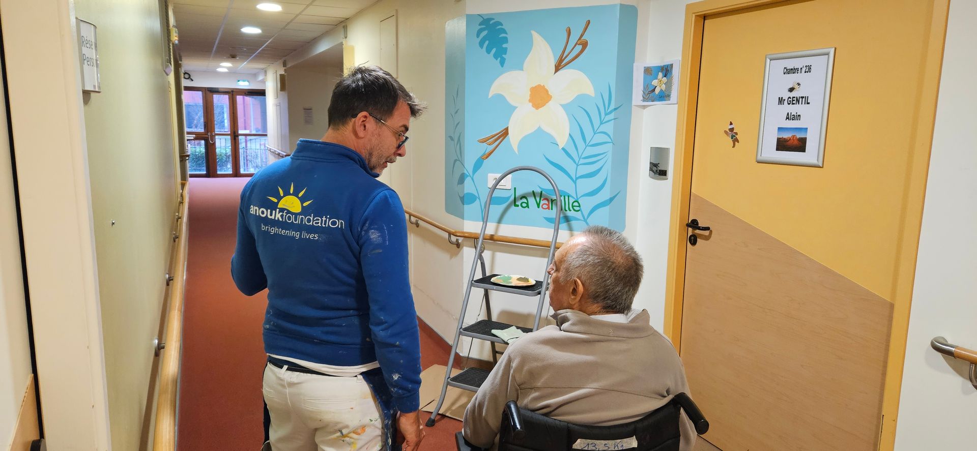 Artist discussing with a seated man in a wheelchair in a hospital hallway next to the mural he is painting
