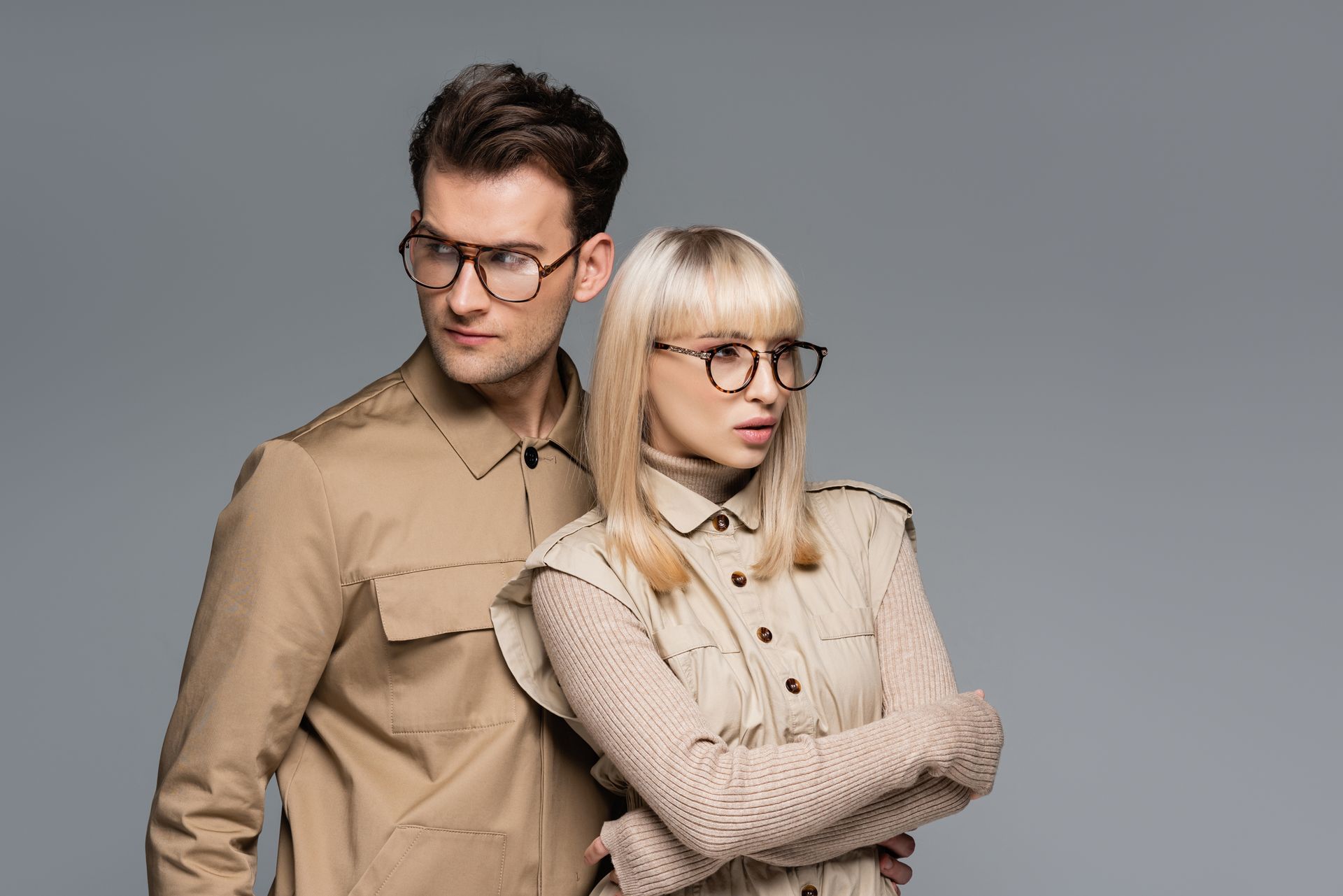 Two stylish models posing against a gray studio background, wearing beige outfits and glasses