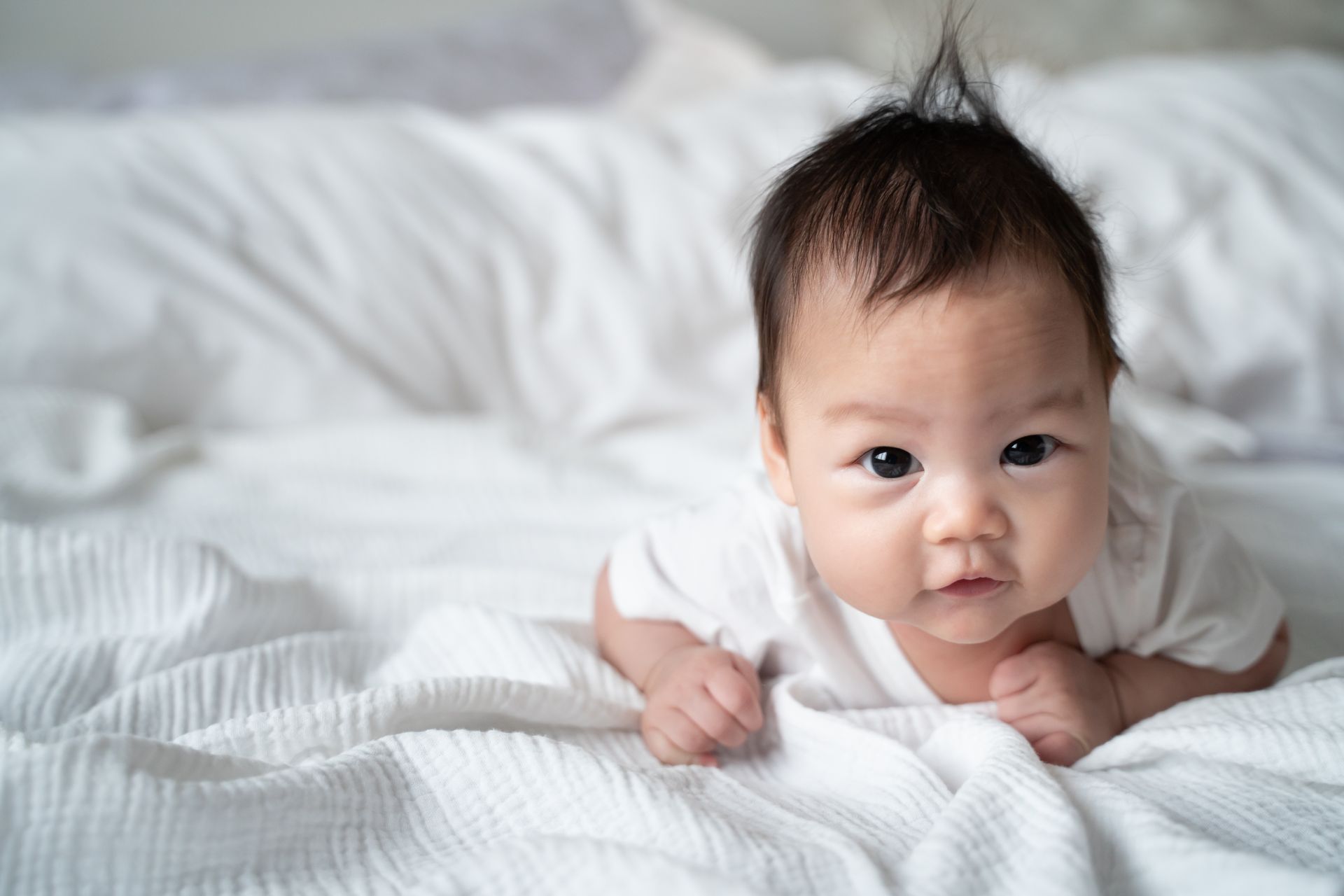A baby is laying on a bed with white sheets and looking at the camera.