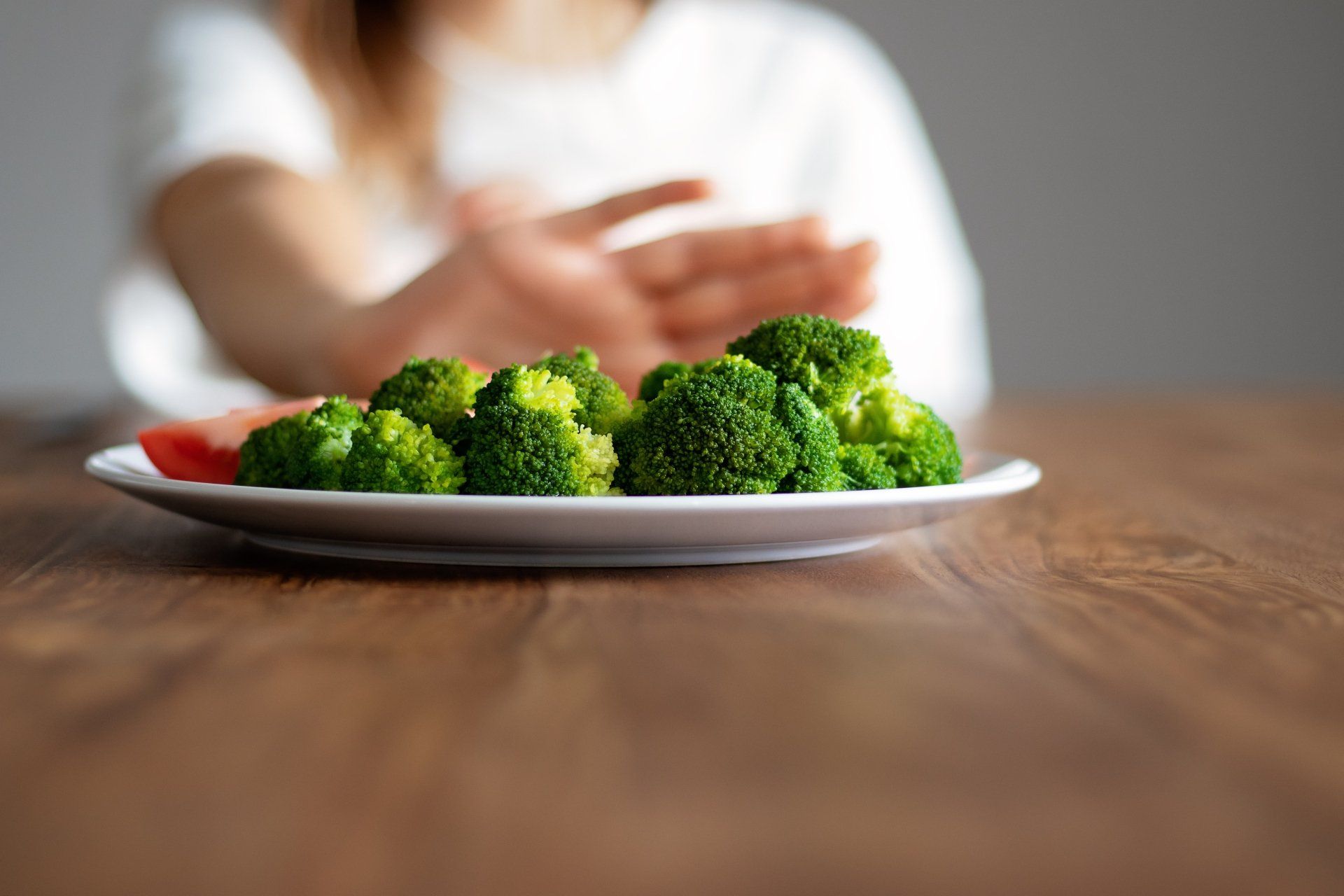 A woman is refusing a plate of broccoli on a wooden table.