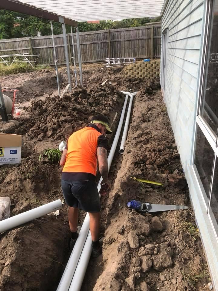 A Man Is Working on A Pipe in The Dirt Next to A House — Polson Construction in Kedron, QLD