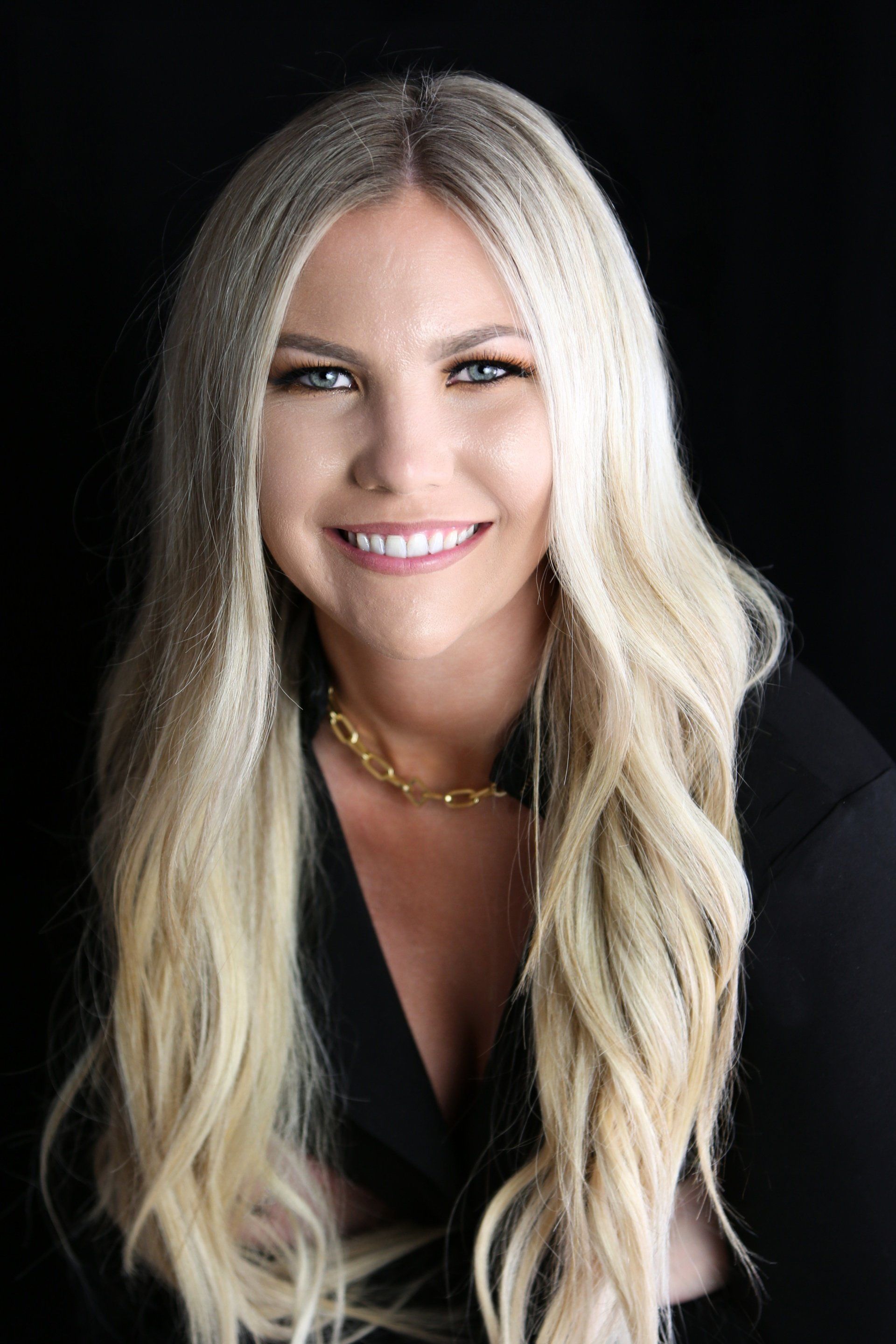 A woman with long blonde hair is wearing a black jacket and smiling.