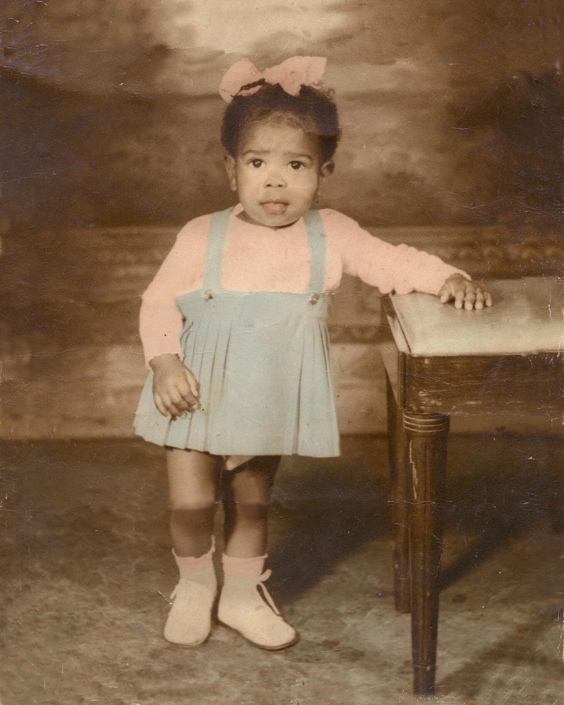 A little girl with a pink bow in her hair is standing next to a table.