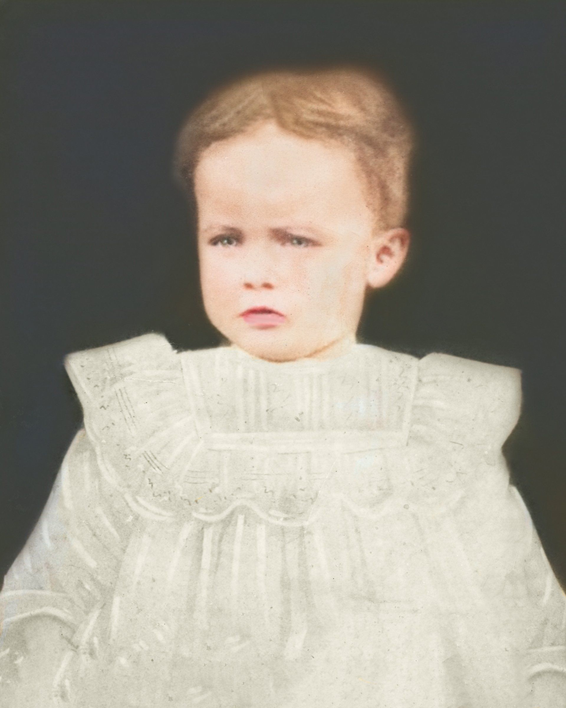 A little boy in a white dress is standing in front of a black background.