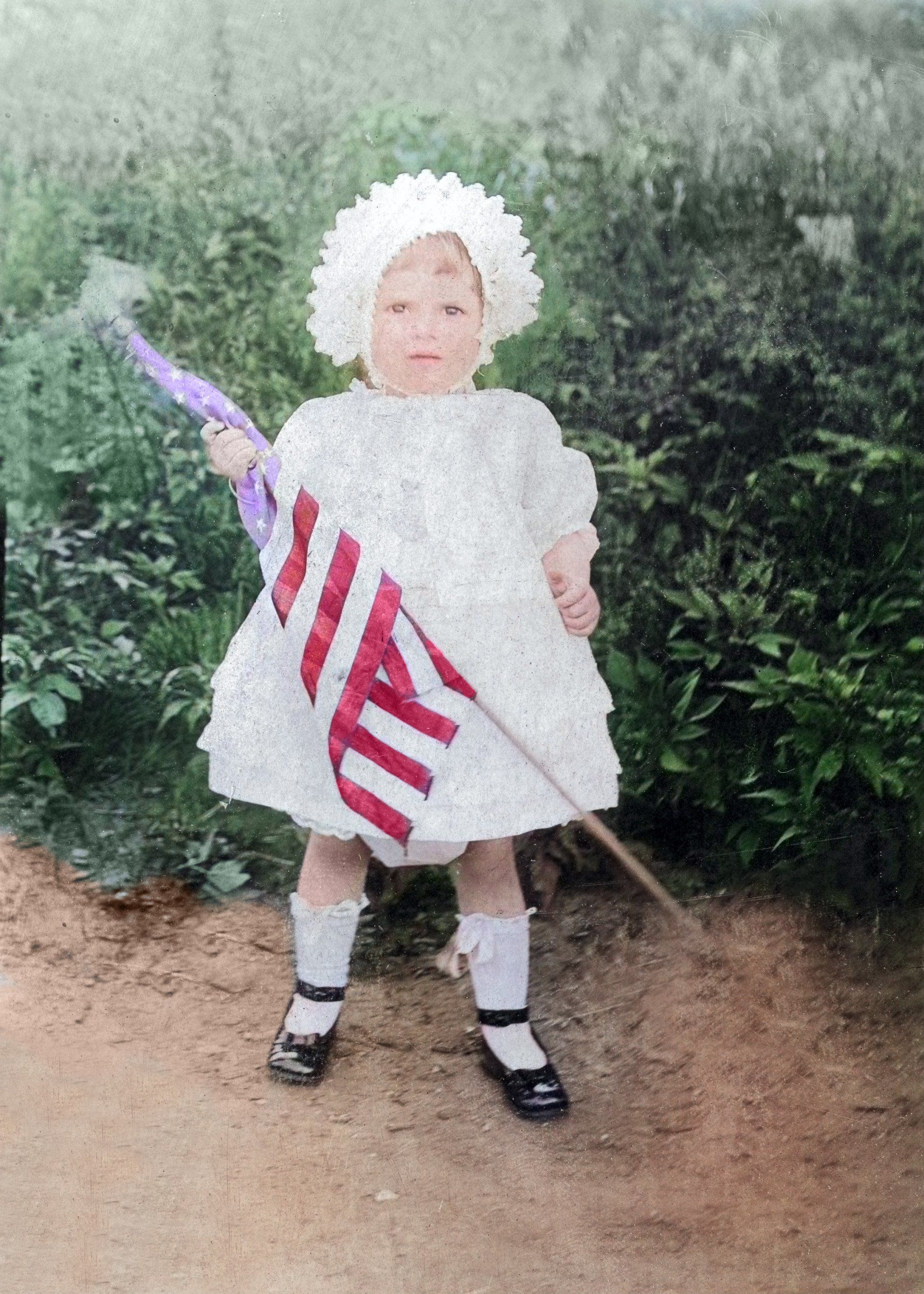 A little girl in a white dress is holding an american flag and an umbrella.
