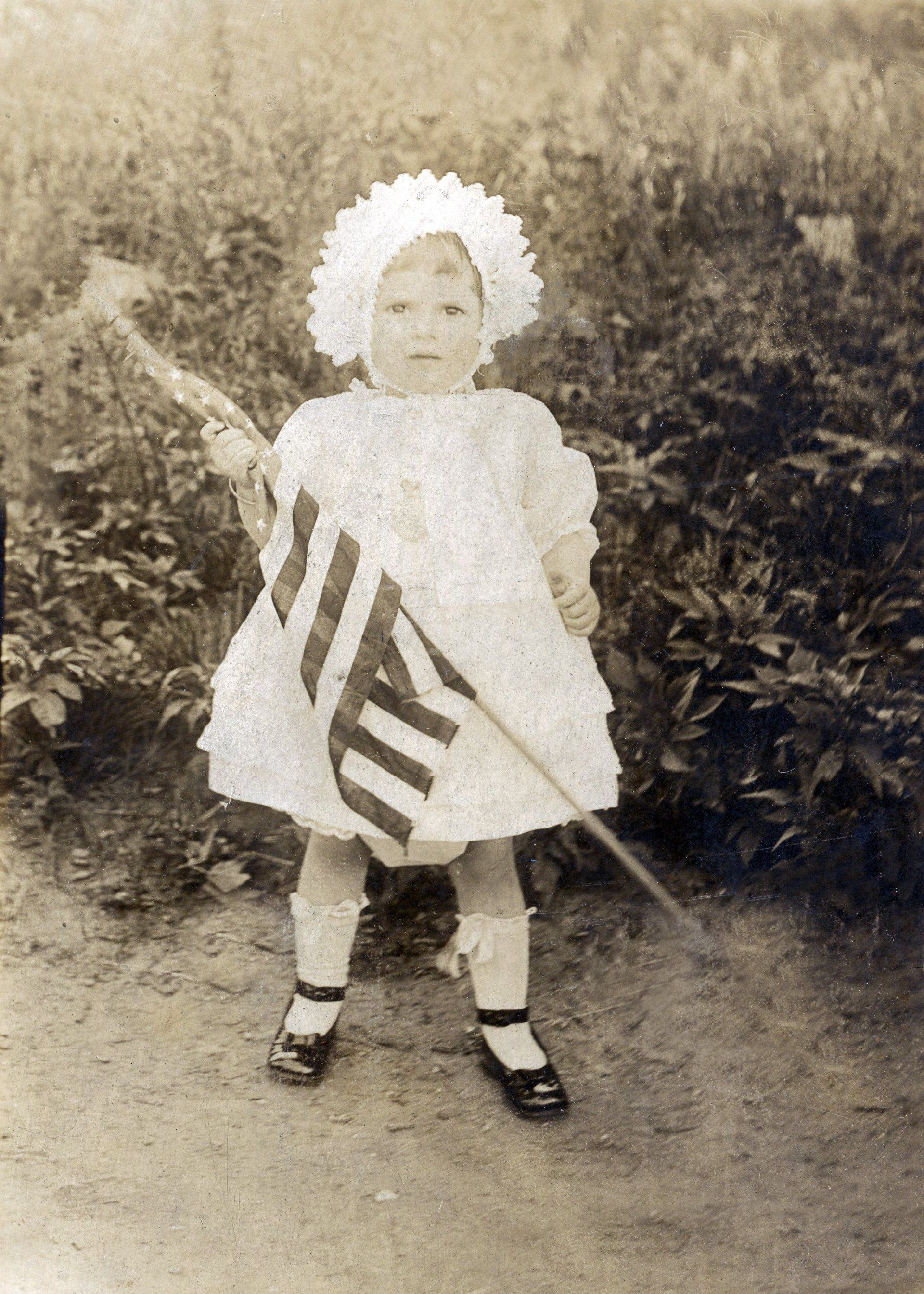 A little girl in a white dress is holding an american flag.