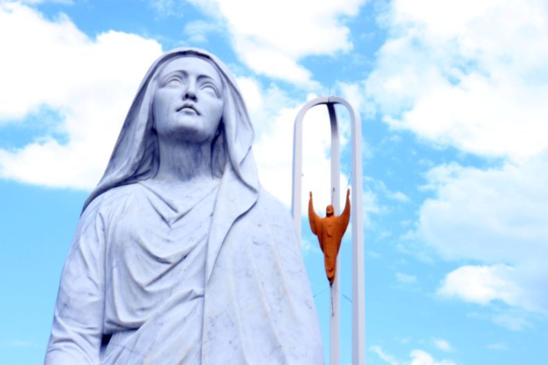 A white statue of a woman looking upward beside an orange sculpture of a figure with outstretched arms against a blue sky.