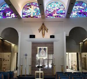 Inside a church, a golden statue of Jesus hangs above an altar with a framed image, framed by colorful stained glass.