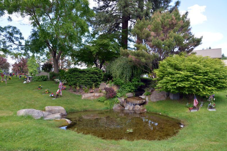 A small pond with a stone waterfall feature nestled in a grassy landscape with mature trees and cemetery markers.