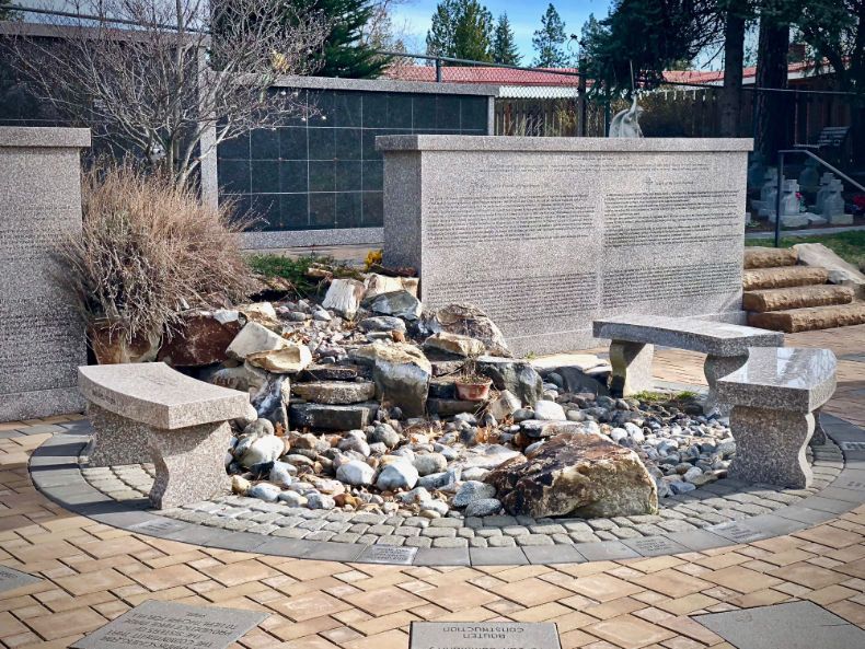 A rock garden with a small waterfall feature, flanked by two stone benches in a paved memorial setting.
