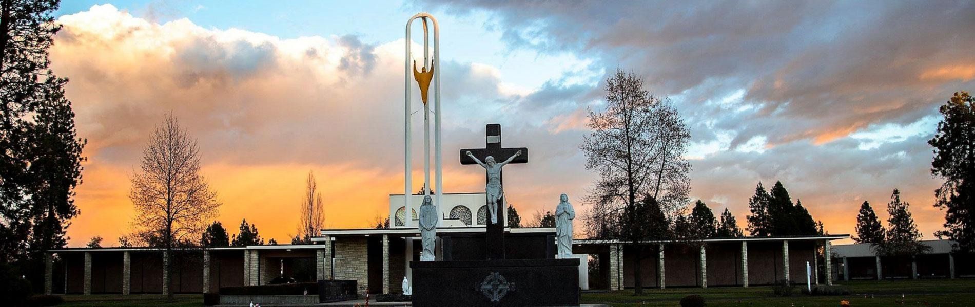 A dark crucifix stands before a modern cemetery chapel under a golden sunset sky.