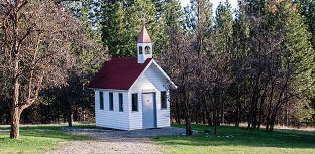 A small, white wooden chapel with a red roof and a bell tower stands in a clearing surrounded by trees.