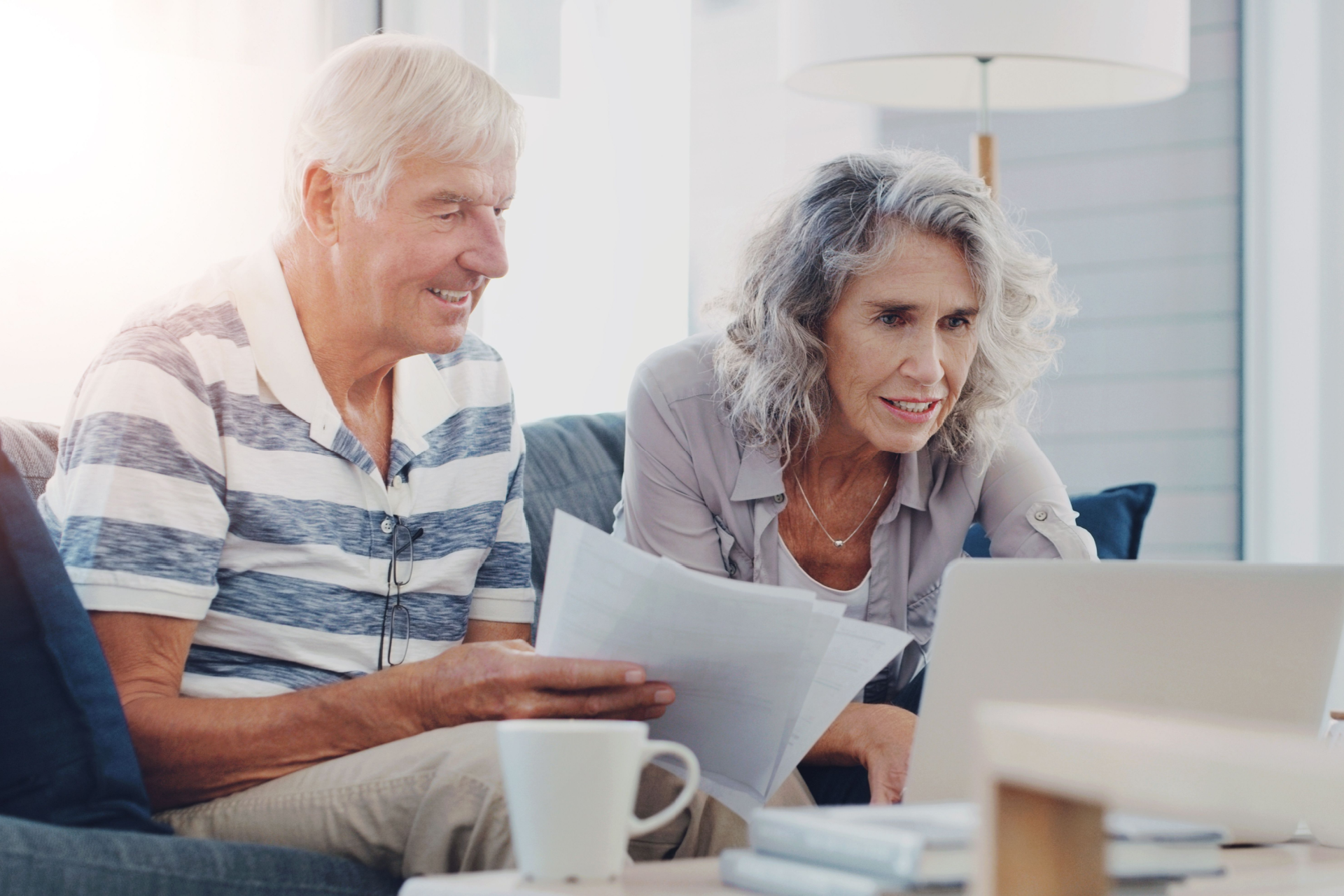 Two people sitting on a sofa and looking at a laptop while holding papers in a bright living room.