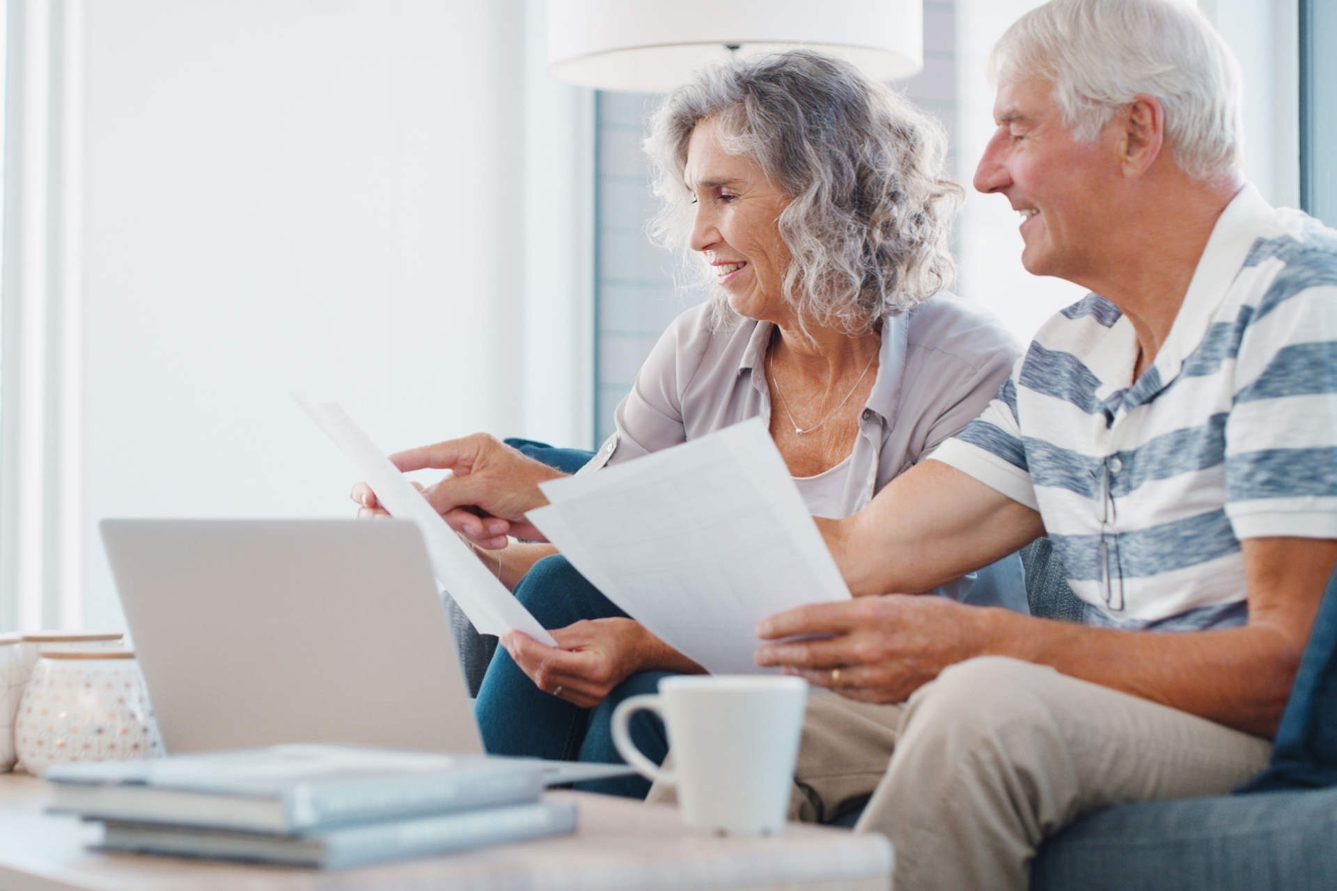 Two people sitting on a sofa, looking at documents and a laptop on a coffee table while smiling.