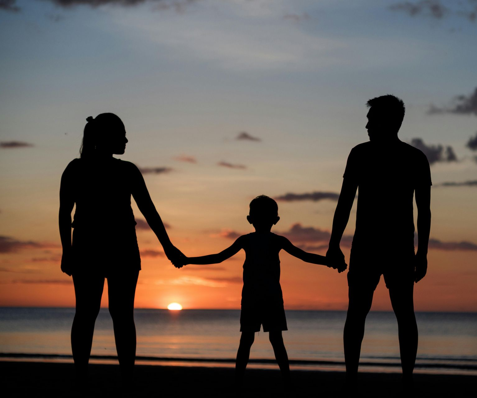 A family silhouetted against a vibrant sunset over the ocean, standing on a beach and holding hands.