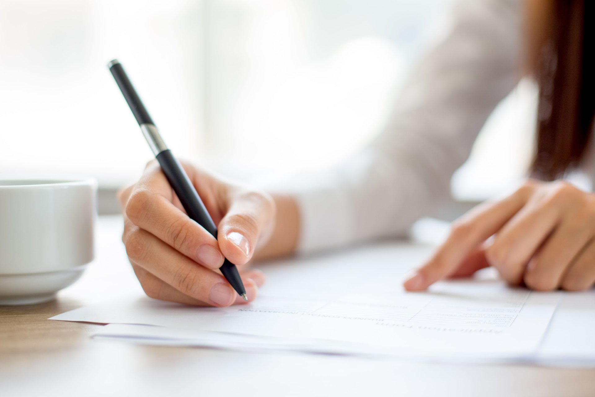 A person's hand holding a pen and writing on a white paper next to a ceramic cup.