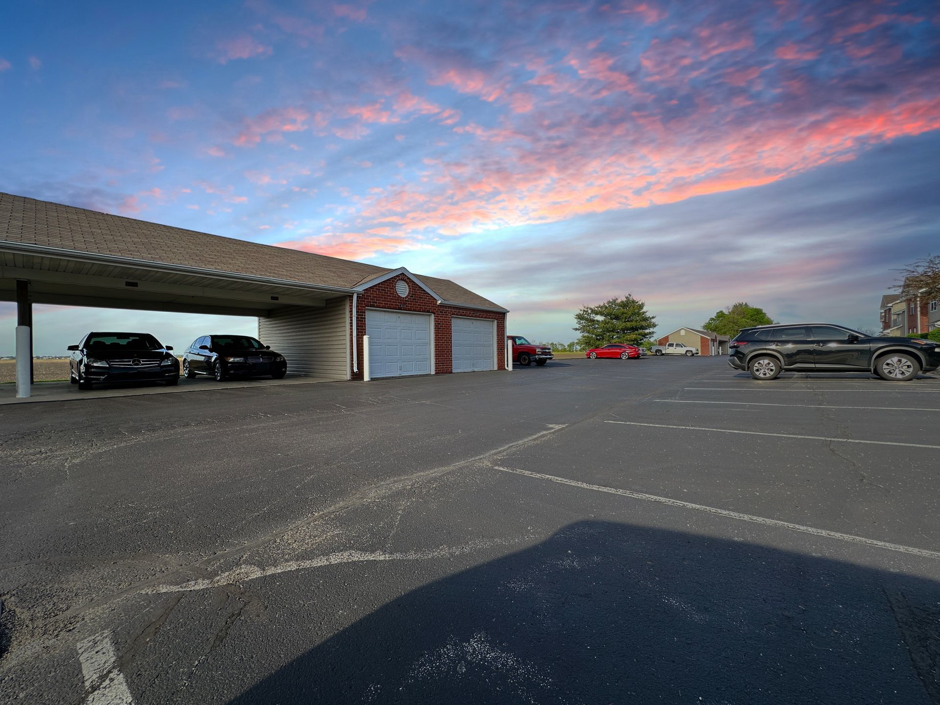 Cars parked under a carport and in a lot, with a brick building in the background, under a colorful sky.