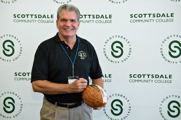 A man is holding a football in front of a wall that says scottsdale community college