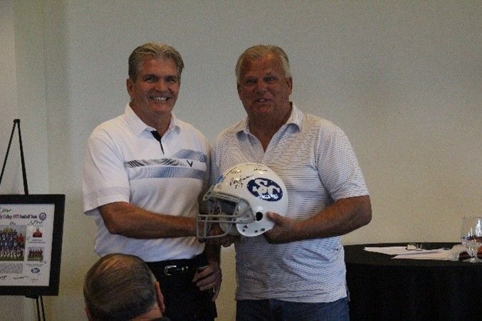 Two men are shaking hands while holding a football helmet