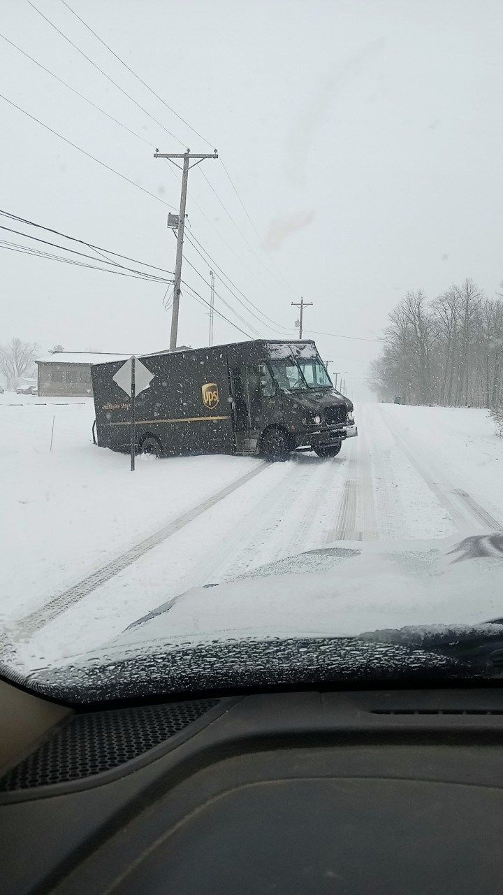 UPS truck stuck in snowy road, surrounded by snow and power lines. The windshield of a vehicle is in the foreground.