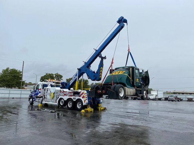 A large tow truck lifting a green semi-truck in a wet, paved area. Two workers are on the ground.
