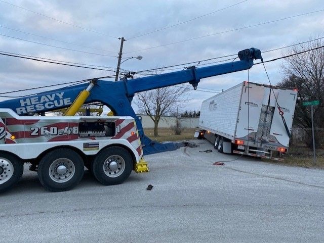 A large tow truck lifts a semi-trailer off the side of a road. The tow truck is blue and white.