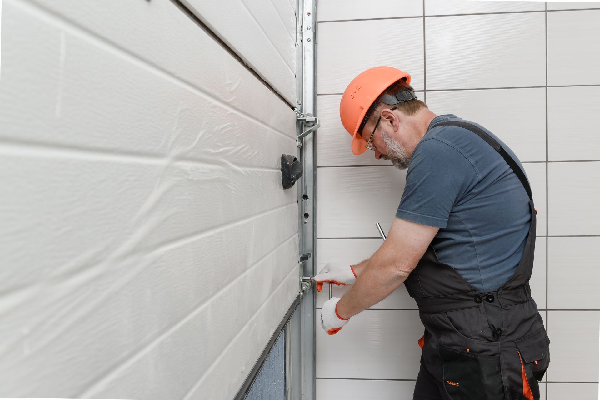 A person in overalls and a hard hat repairs a garage door with tools.