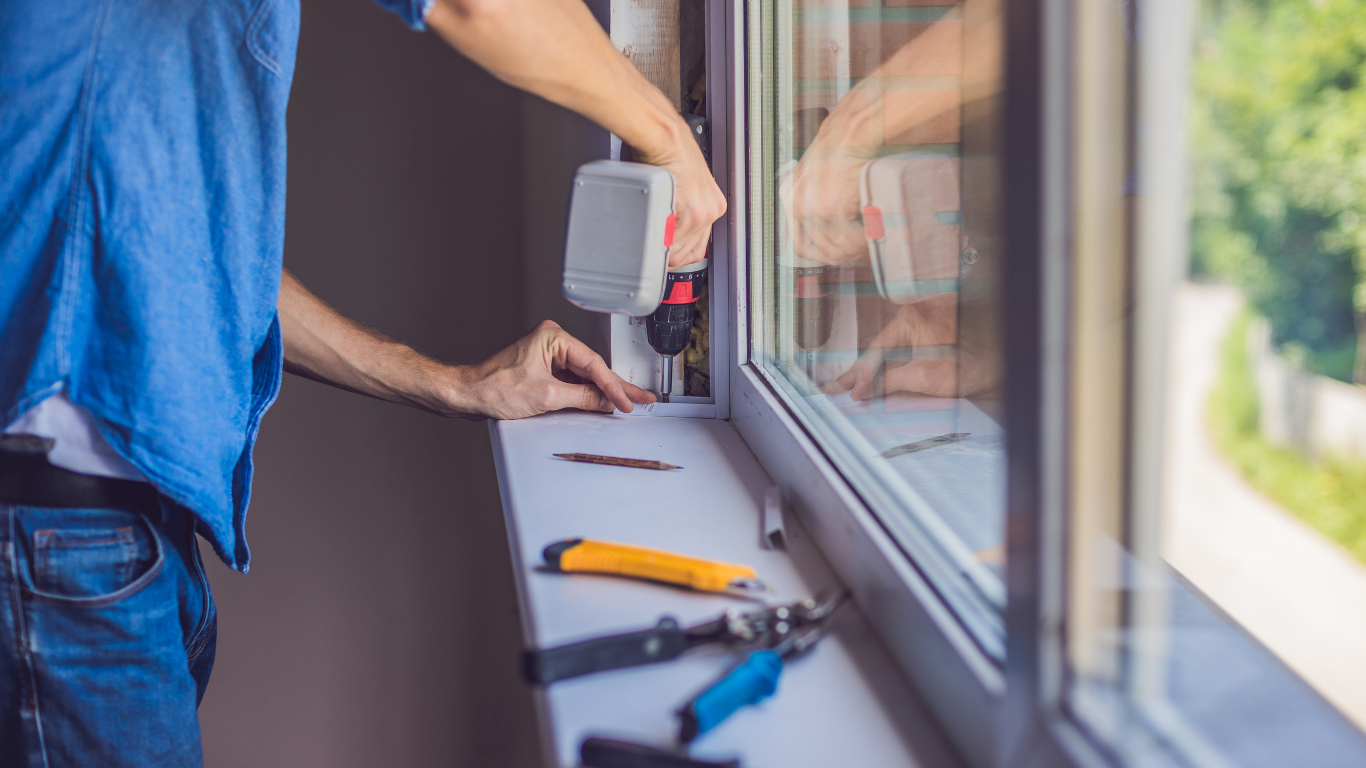 Person in blue shirt using a power drill on a window frame; tools laid out on the windowsill.