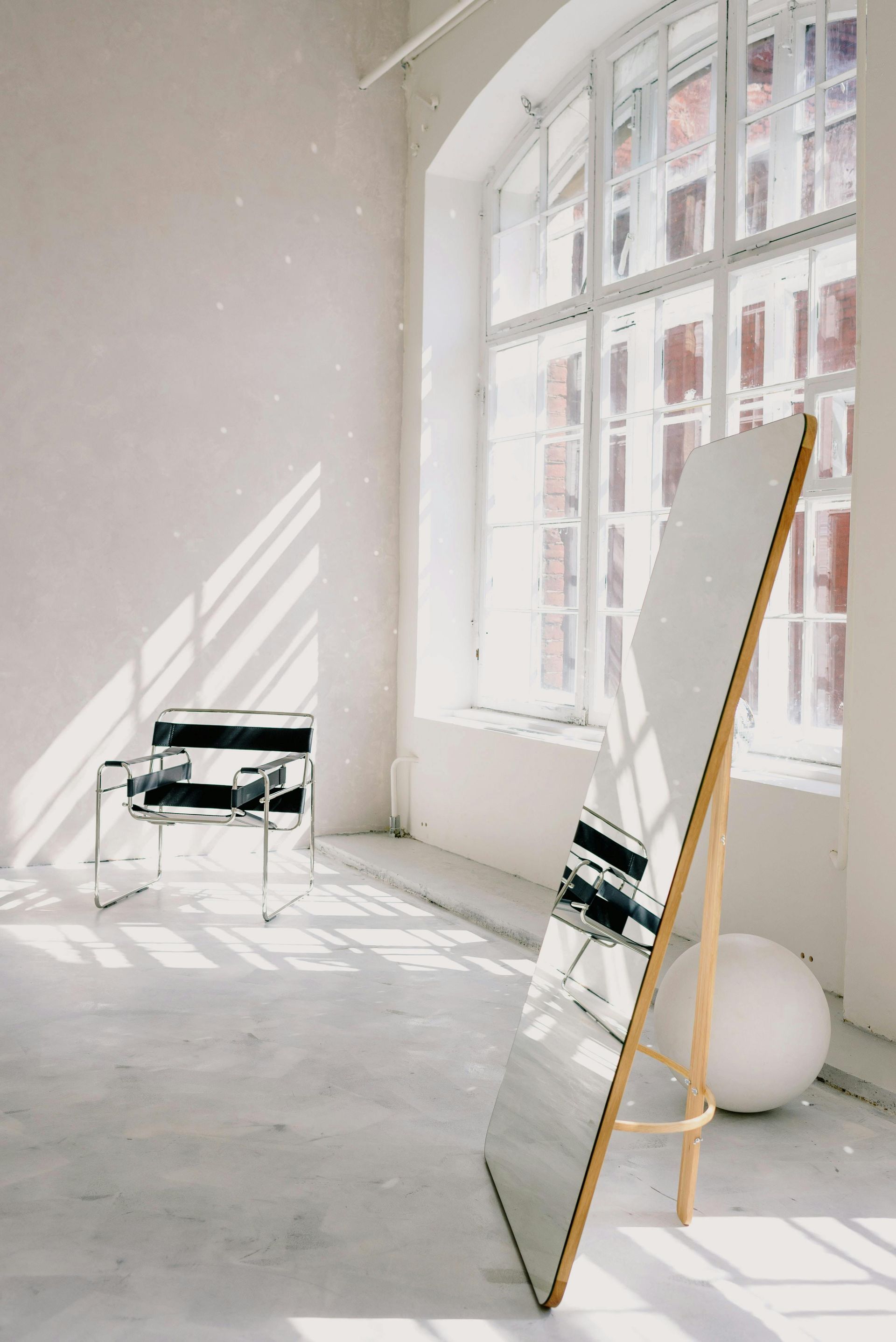 White room with large mirror, window, chair, and sphere, casting sunlight shadows.