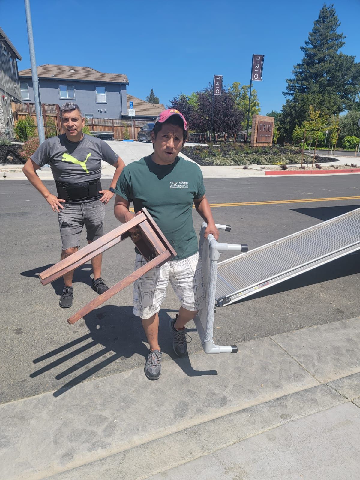 Two people stand outdoors on an asphalt street, one holding a wooden frame and the other a folding table or ramp.