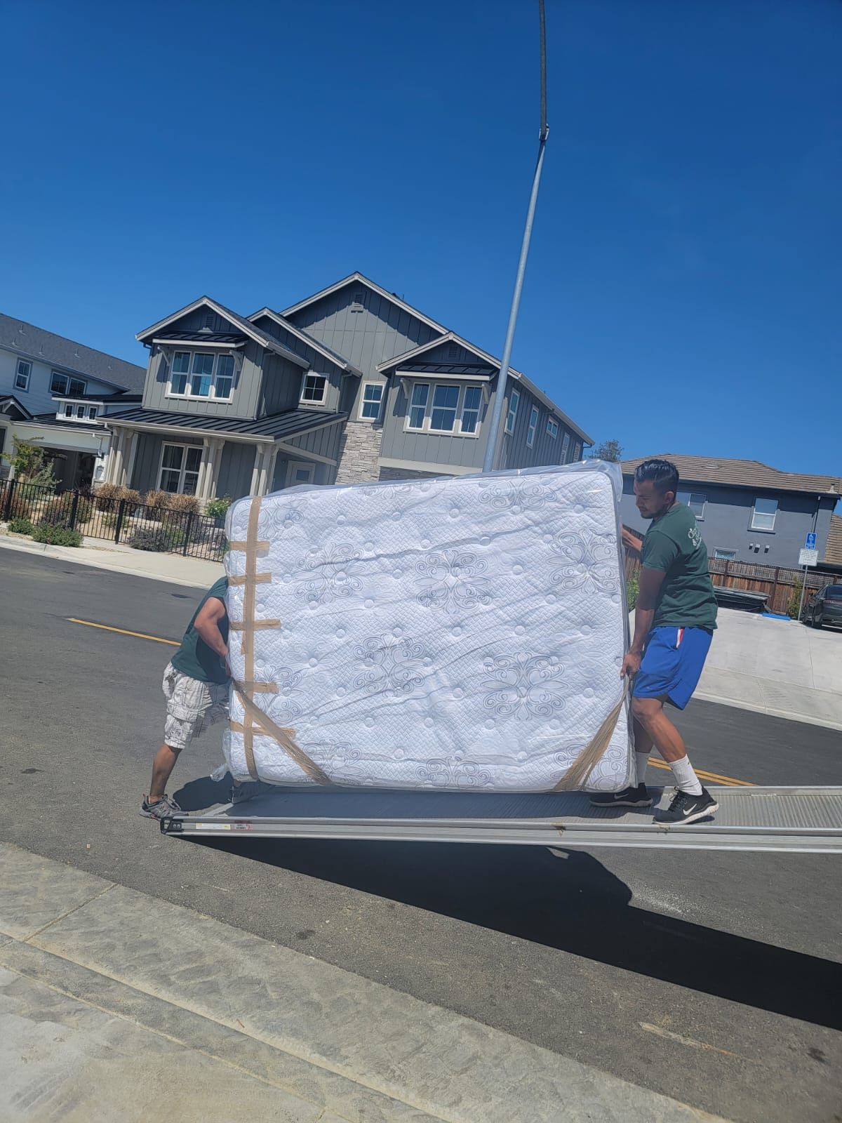 Two people load a large, plastic-wrapped white mattress onto a truck lift gate in a suburban neighborhood.