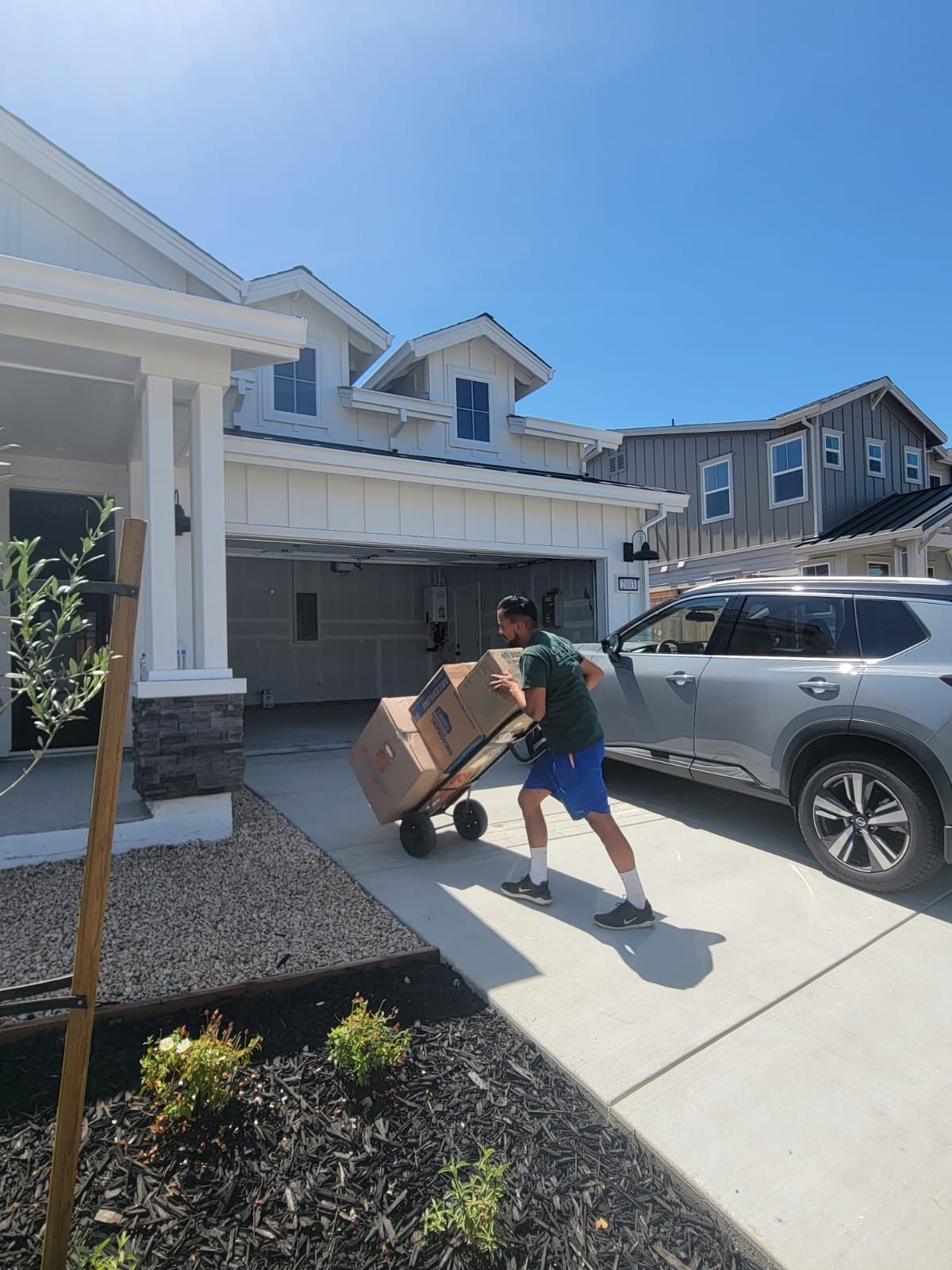 A person in a green shirt and blue shorts pulls a hand truck loaded with cardboard boxes toward a suburban house garage.