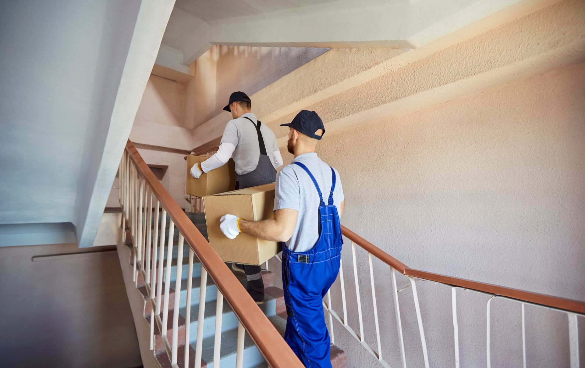 Two professional movers in uniforms carry cardboard moving boxes up a narrow indoor staircase safely