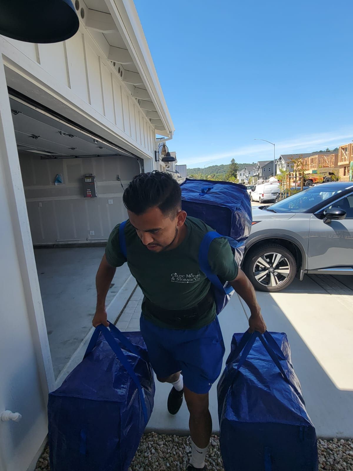 A person carries three large blue moving bags while walking from a residential garage toward a parked car.