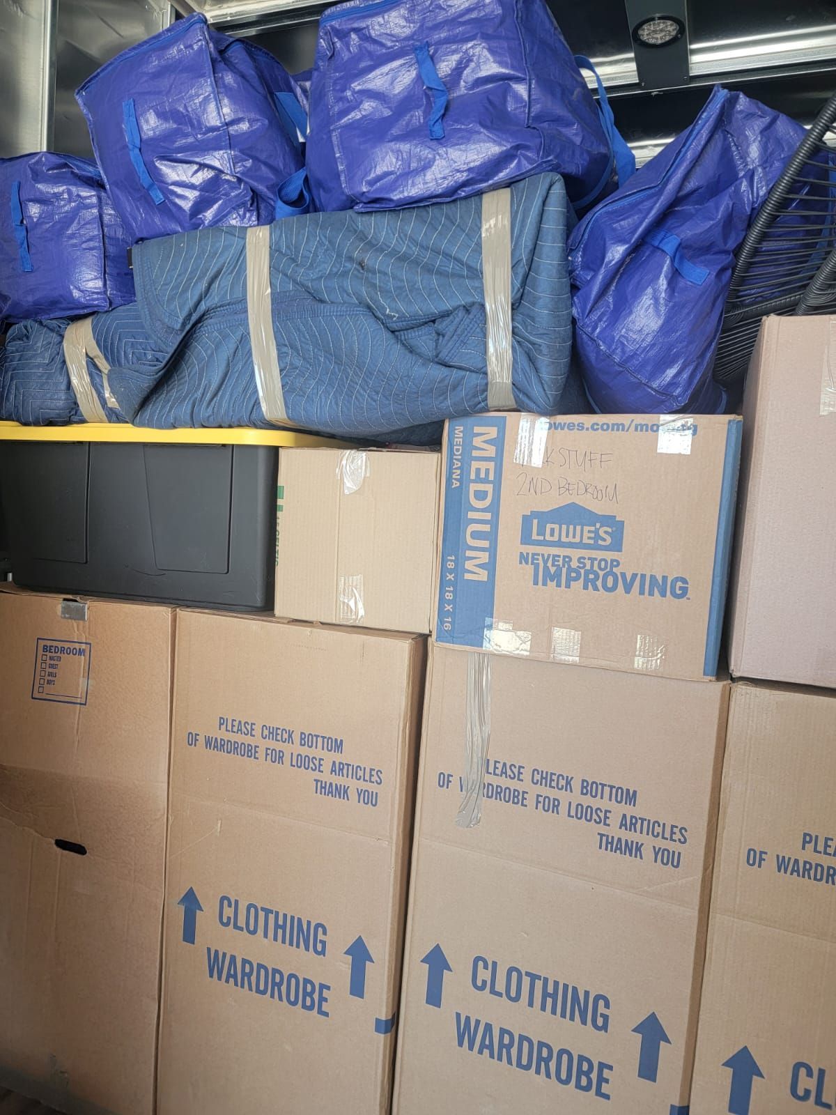 Stacked cardboard moving boxes, blue storage bags, and furniture wrapped in moving blankets inside a moving truck.
