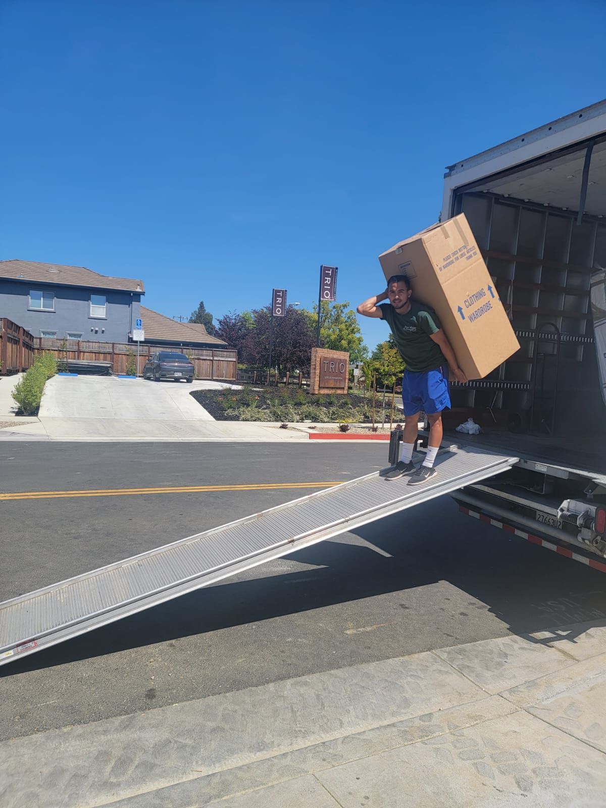 A person carrying a large cardboard box down a metal ramp from the back of a moving truck on a sunny residential street.