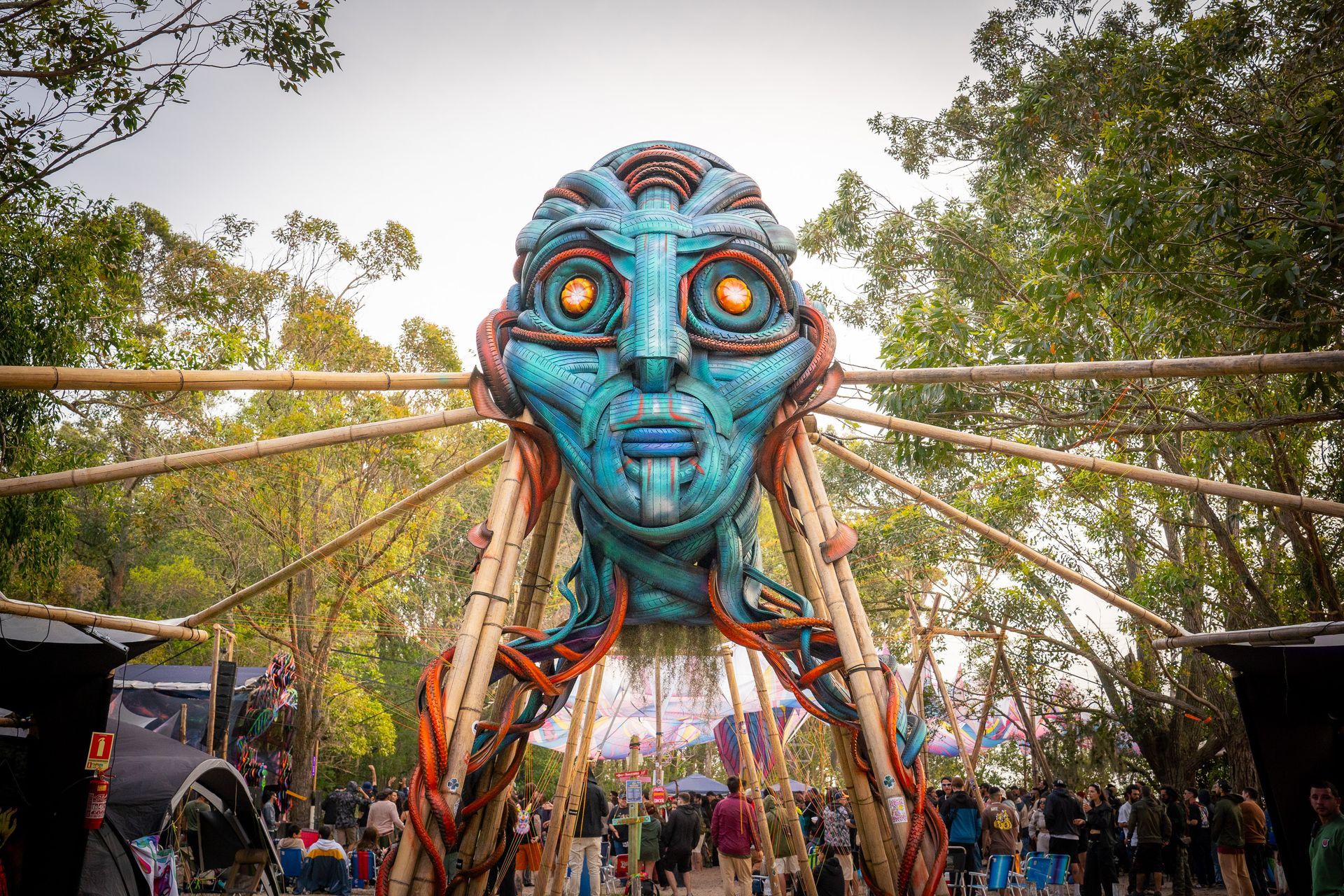 Grande escultura azul de um rosto tribal em um festival ao ar livre, com luzes e multidões embaixo dela.