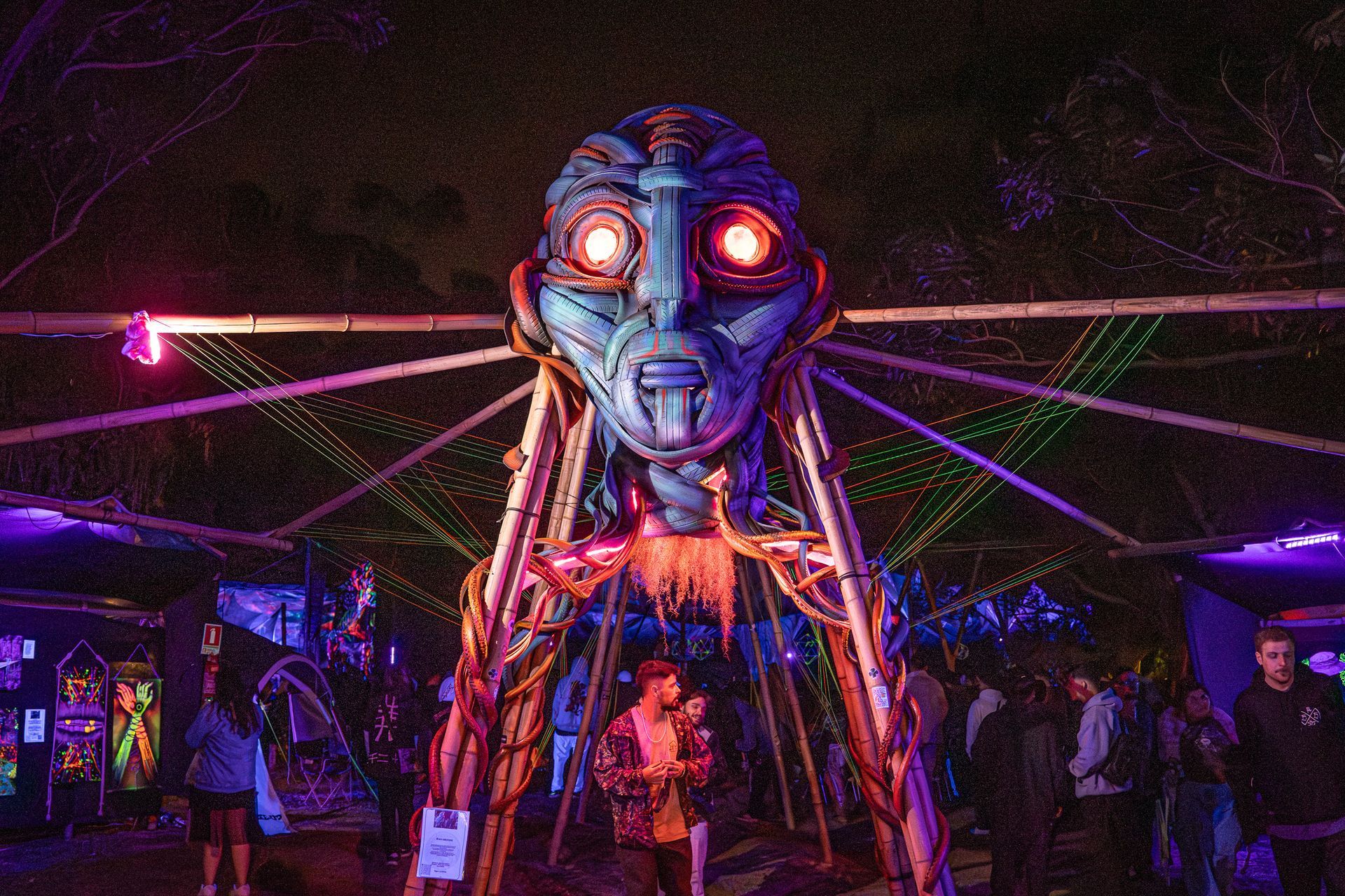 Escultura brilhante de um rosto alienígena com olhos vermelhos em um festival noturno iluminado por luzes de néon.