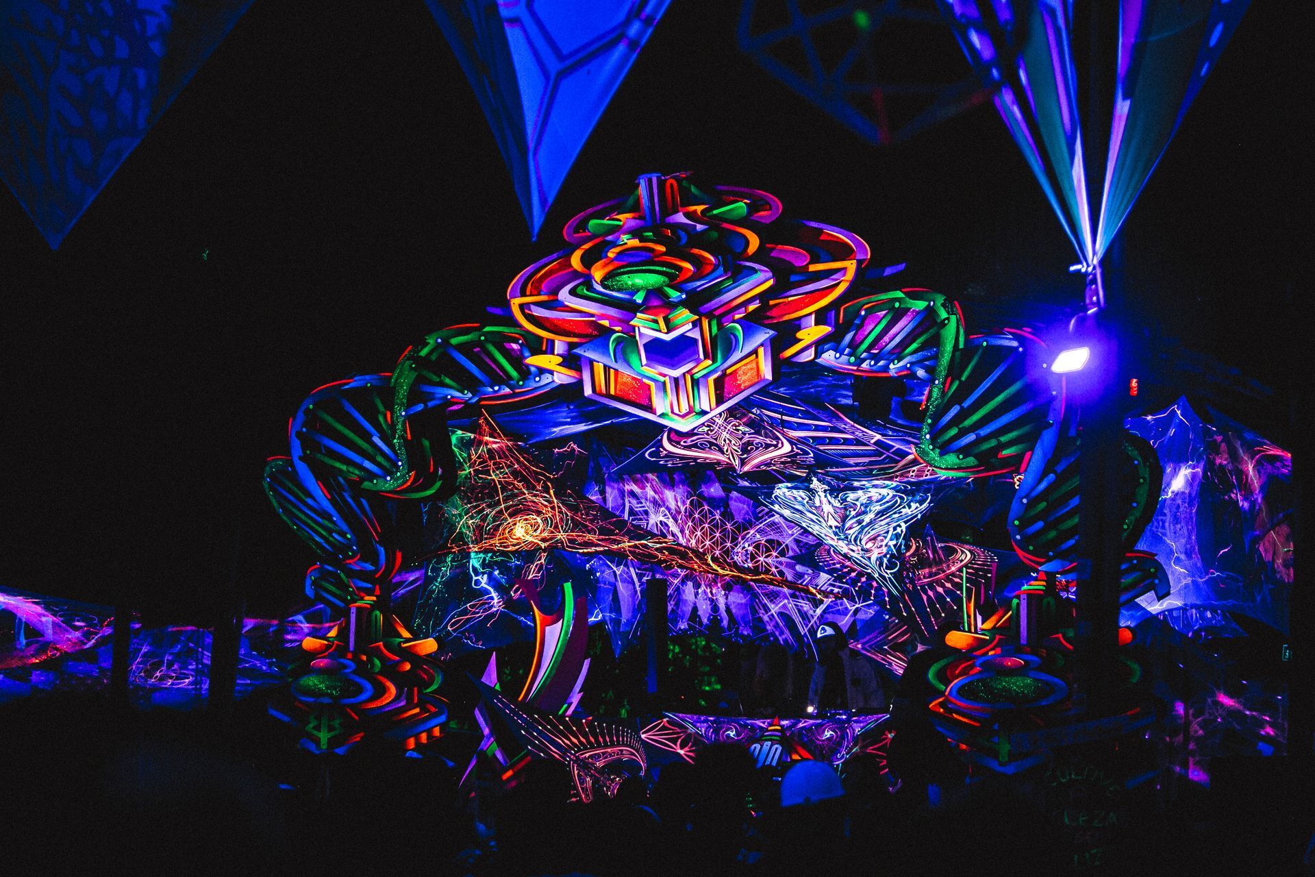 Palco iluminado com luzes de néon, esculturas abstratas brilhantes e luzes laser em um ambiente escuro de concerto.
