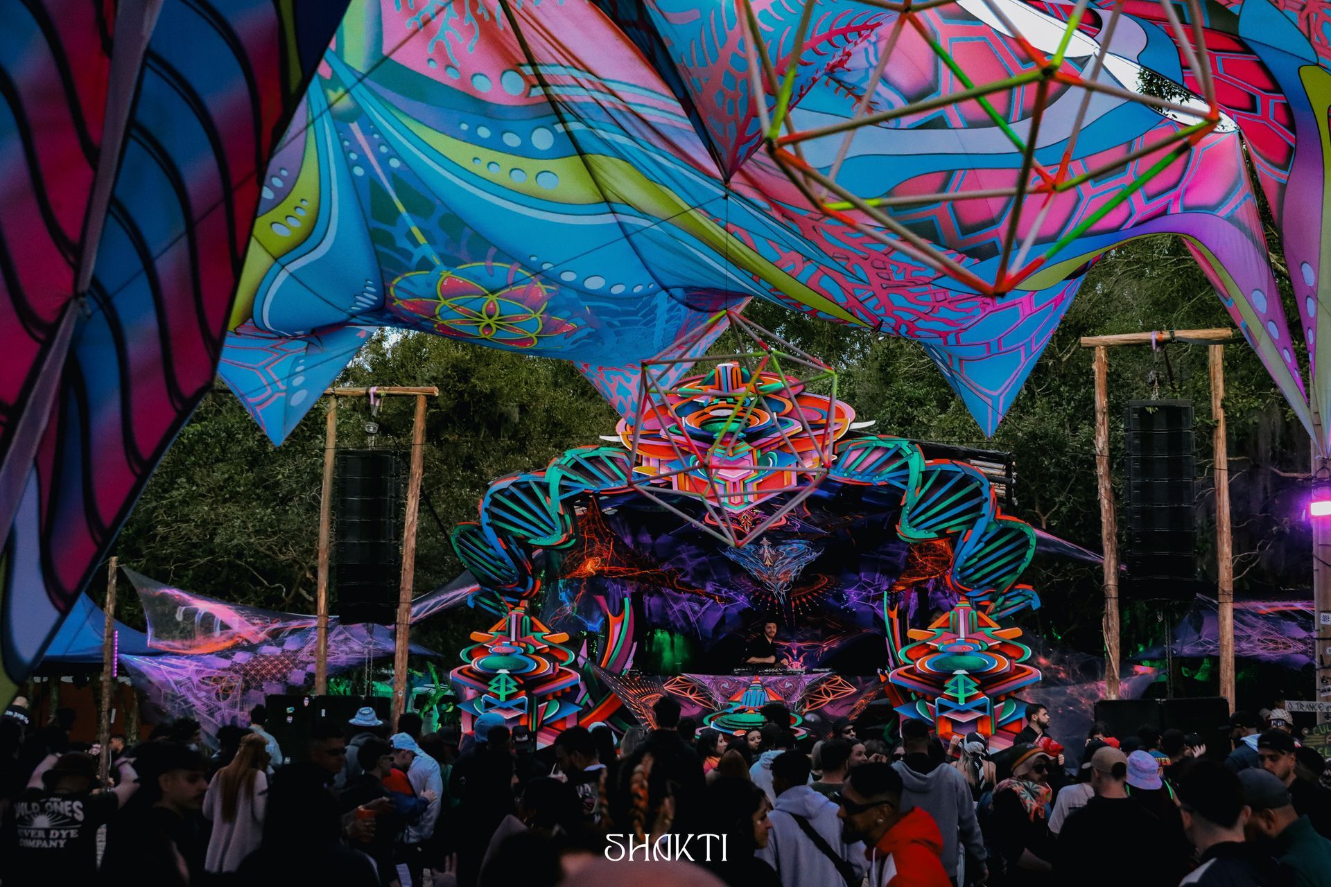 Multidão em um palco colorido de festival ao ar livre sob luzes geométricas de néon.