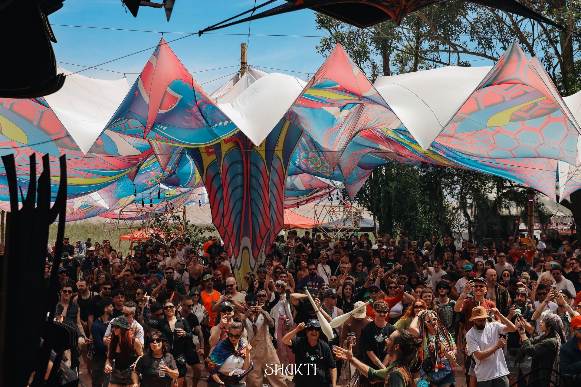 Multidão dançando sob toldos geométricos coloridos em um festival ao ar livre.