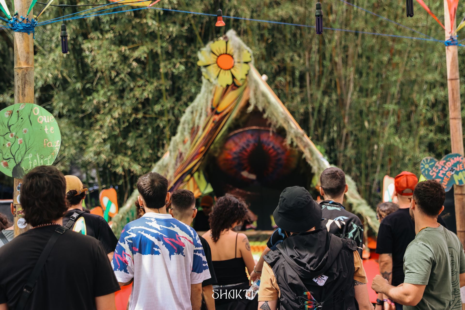 Multidão de frente para um palco colorido ao ar livre com uma grande decoração em forma de sol e um toldo triangular.