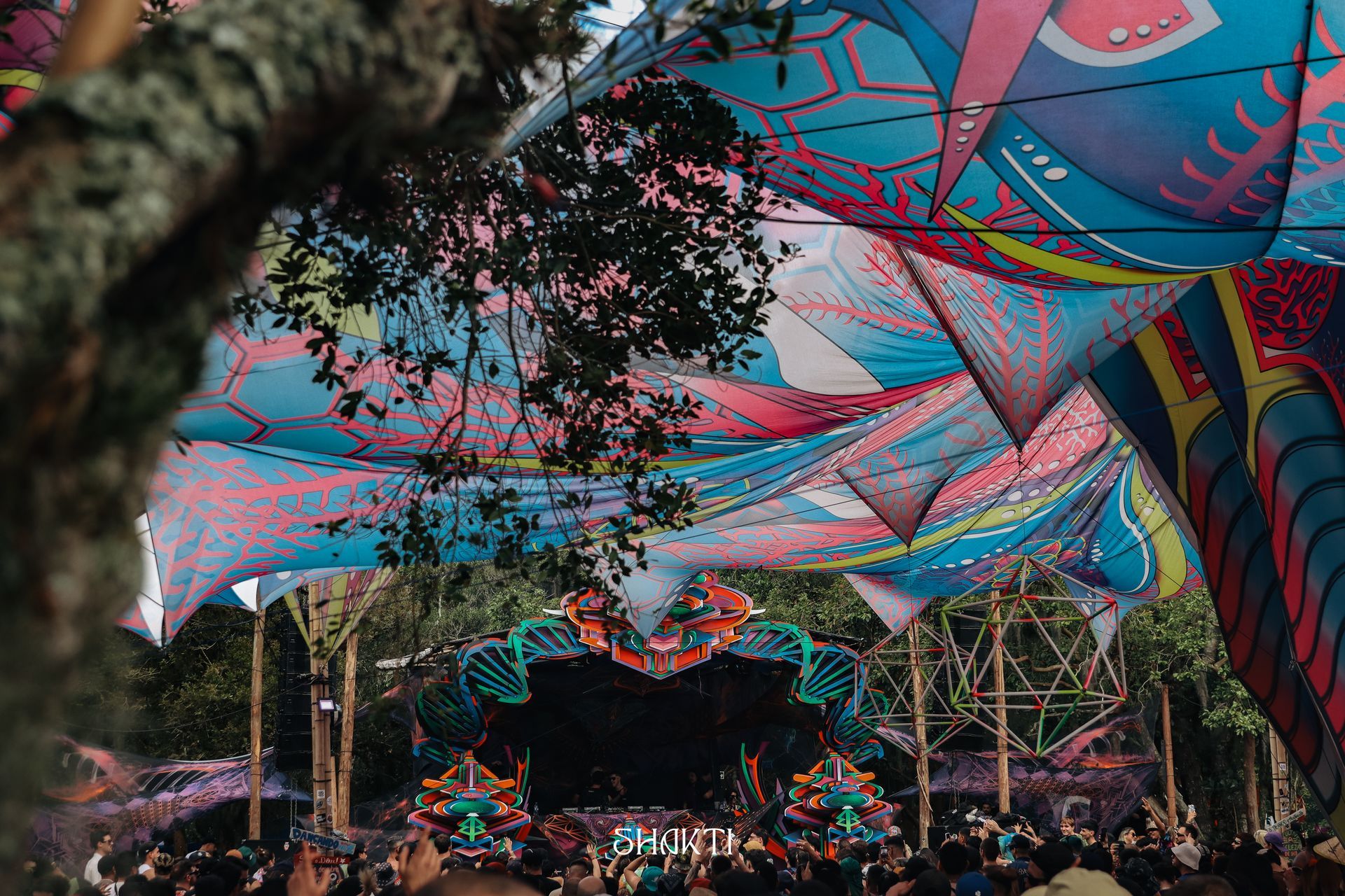 Cobertura geométrica colorida sobre um palco lotado de festival ao ar livre ao entardecer.