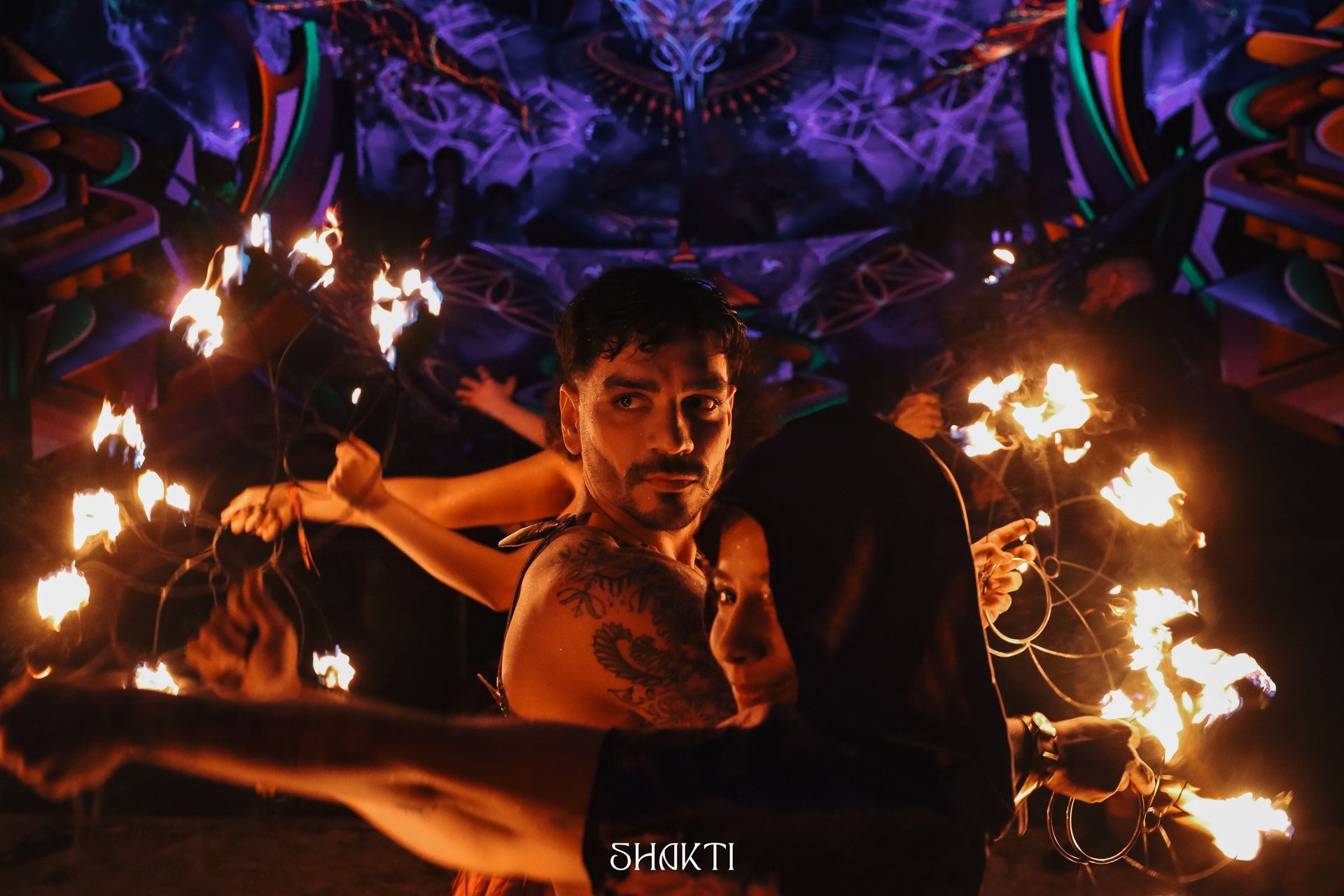 Artista de fogo com bastões em chamas em um cenário escuro e colorido.