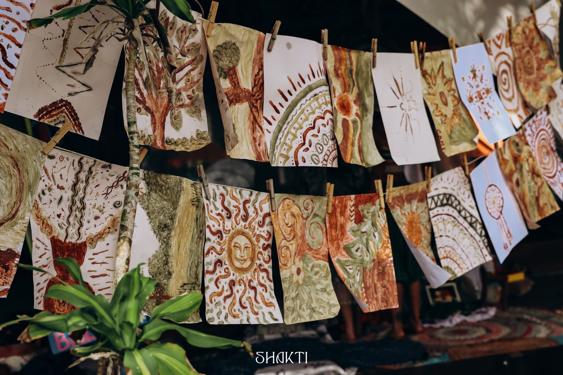 Varal com painéis de tecido pintados à mão em tons terrosos, exposto ao ar livre acima de plantas verdes.