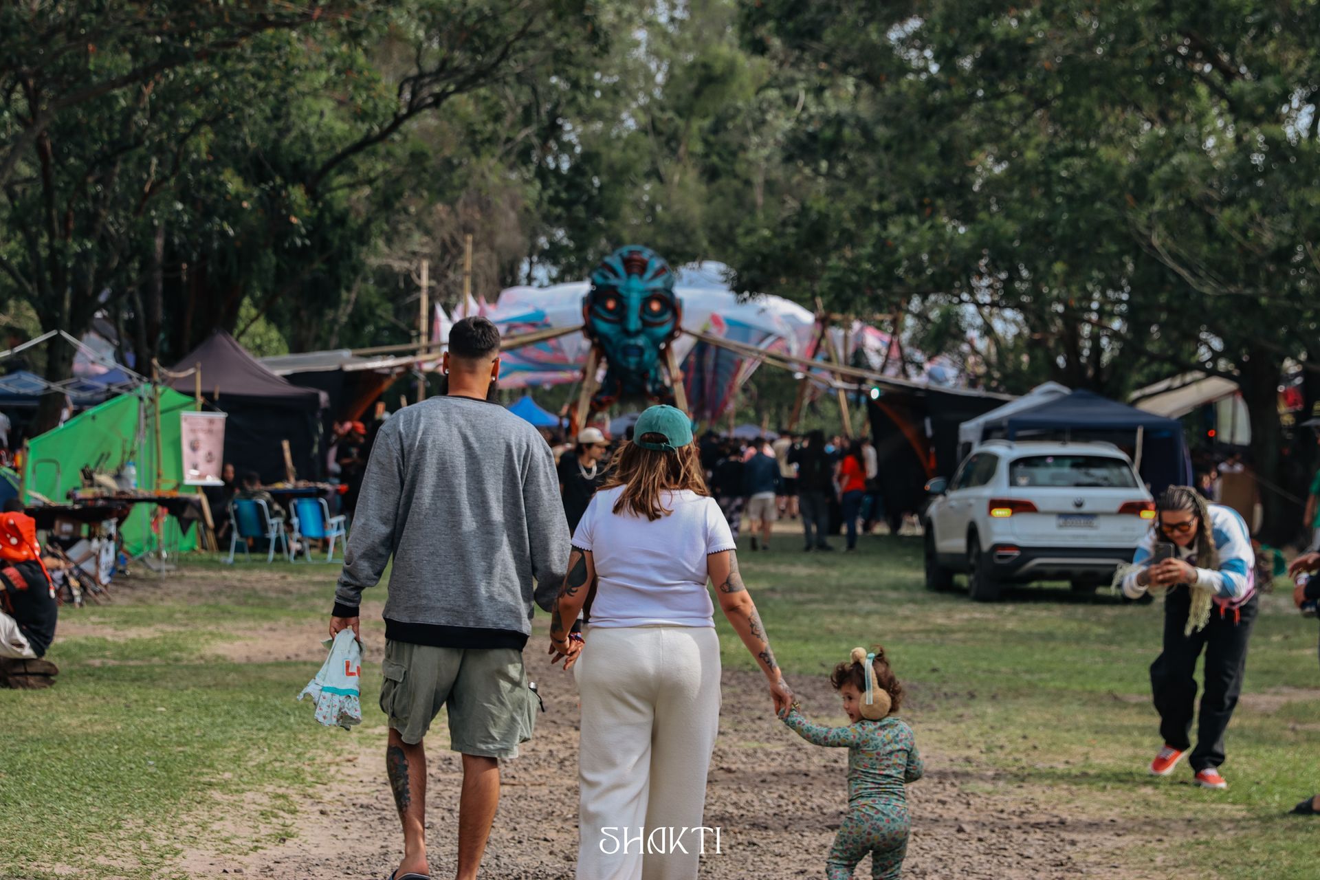 Pessoas caminhando com uma criança em um parque gramado, com barracas, carros e árvores ao fundo.