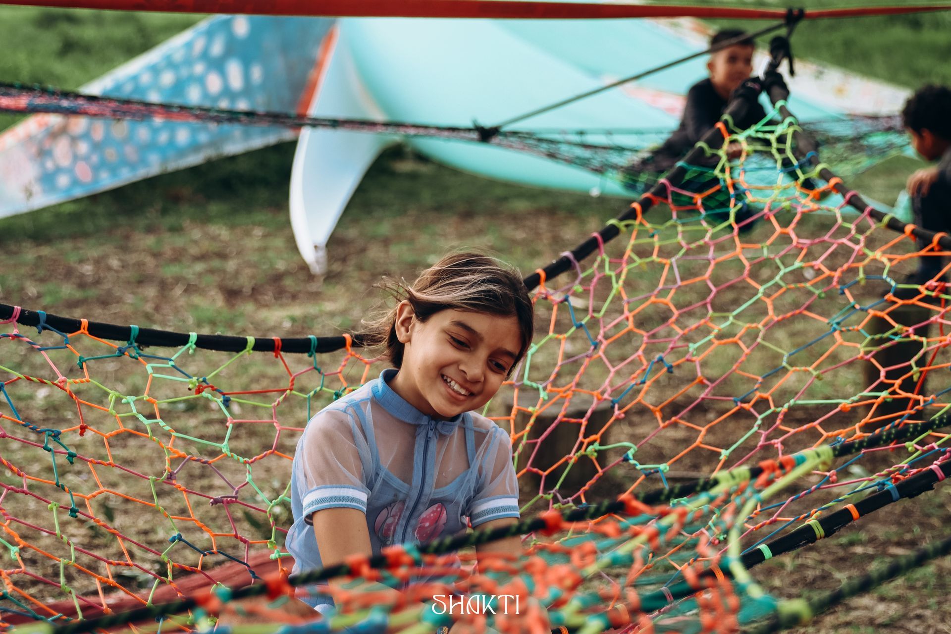 Criança sorrindo em uma rede colorida ao ar livre, com outras pessoas desfocadas ao fundo.